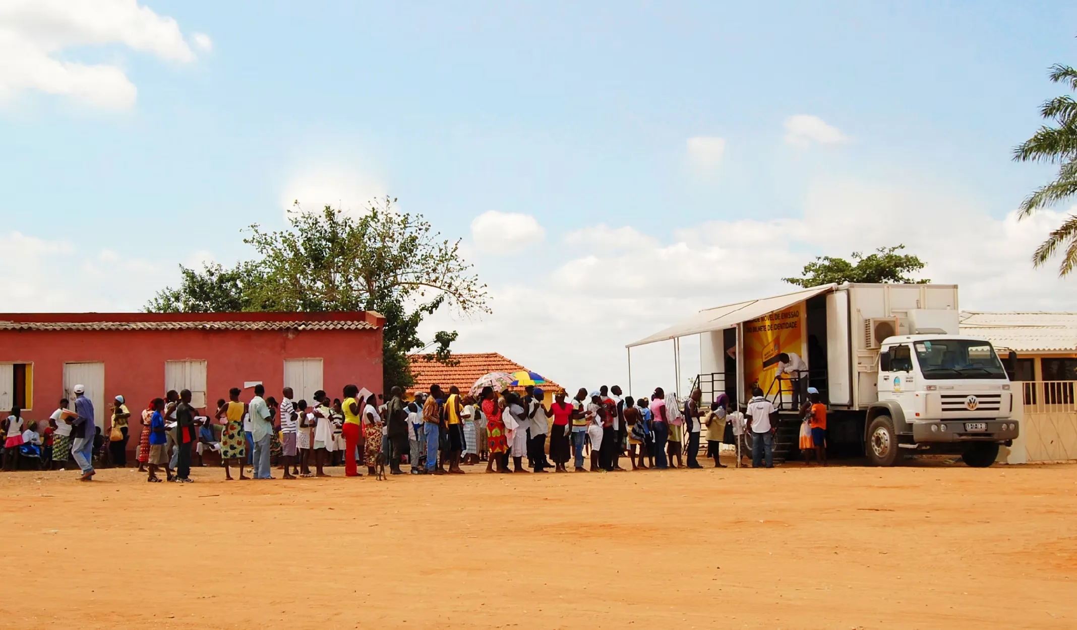 Mobile identity card unit in Angola: a white truck with an awning serves a long queue of residents in an open dirt square, among low houses, under an open sky.